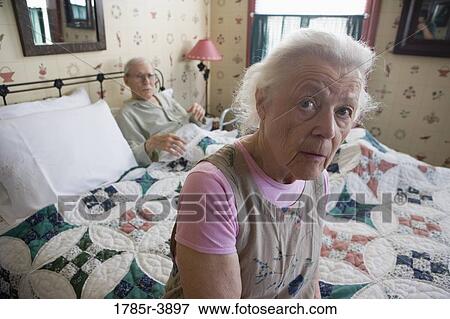 Stock Photo - Worried senior woman sitting on bed with bedridden senior man. Fotosearch