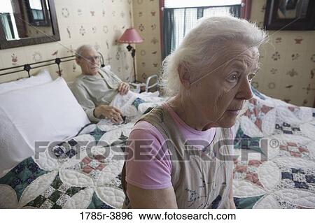 Stock Photograph - Worried senior woman sitting on bed with bedridden senior man. Fotosearch