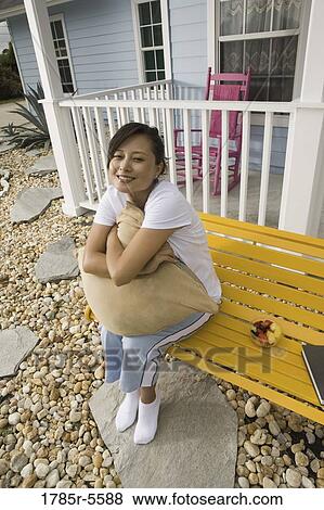 Young Asian woman sitting on a bench hugging a pillow in front of house View Large Photo Image Stock Photo - Young Asian woman sitting on a bench hugging a pillow in front of house. Fotosearch