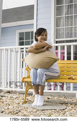 Stock Photo - Young Asian woman sitting on a bench hugging a pillow in front of house. Fotosearch