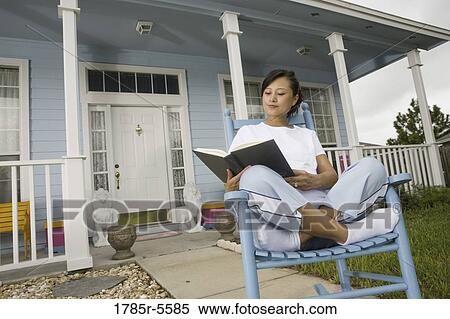 Stock Photography - Young Asian woman sitting on a rocking chair reading a book in front of house. Fotosearch