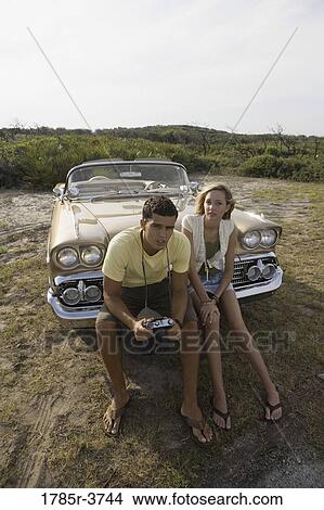 Picture - Young couple sitting on front bumper of antique convertible car. Fotosearch