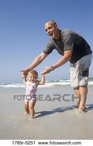 Stock Image - Young father holding baby girl by the hands, helping her walk on the beach. Fotosearch