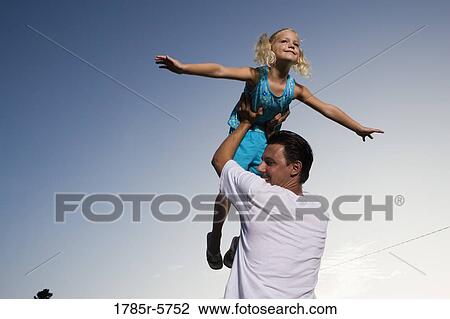 Stock Image - Young girl with arms outstretched, lifted by her father. Fotosearch