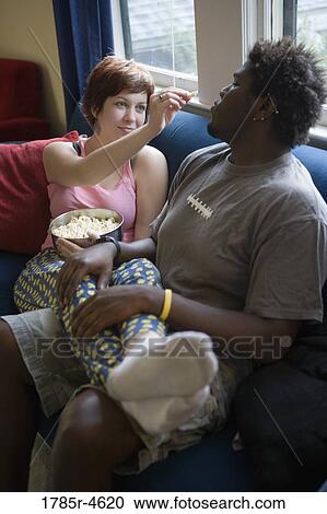 Stock Image - Young inter-racial couple sitting on a sofa eating popcorn. Fotosearch