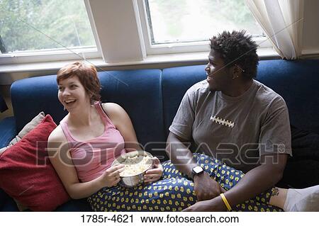 Stock Image - Young inter-racial couple sitting on a sofa eating popcorn. Fotosearch
