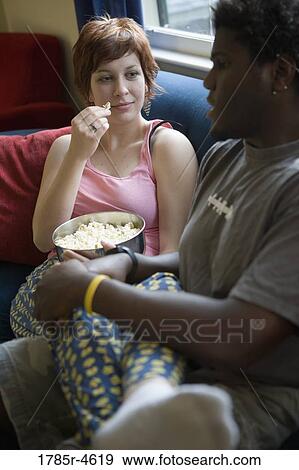 Stock Photo - Young inter-racial couple sitting on a sofa eating popcorn. Fotosearch