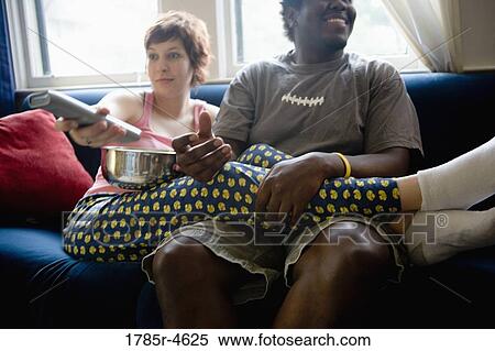 Young inter-racial couple sitting on a sofa watching television View Large Photo Image Stock Photography - Young inter-racial couple sitting on a sofa watching television. Fotosearch