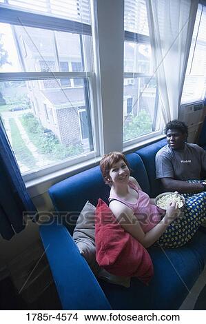 Picture - Young inter-racial couple sitting on sofa eating popcorn. Fotosearch