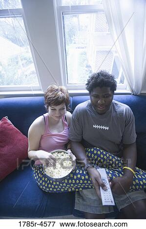 Young inter-racial couple sitting on sofa eating popcorn and watching television View Large Photo Image Stock Photo - Young inter-racial couple sitting on sofa eating popcorn and watching television. Fotosearch