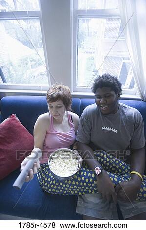 Stock Photo - Young inter-racial couple sitting on sofa eating popcorn and watching television. Fotosearch