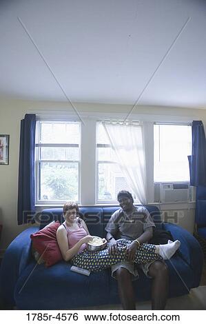 Young inter-racial couple sitting on sofa eating popcorn View Large Photo Image Stock Photograph - Young inter-racial couple sitting on sofa eating popcorn. Fotosearch