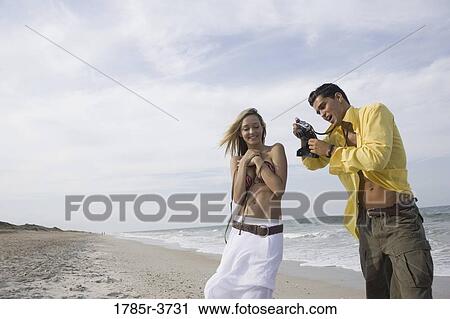 Stock Image - Young man photographing scantily clad woman on the beach. Fotosearch