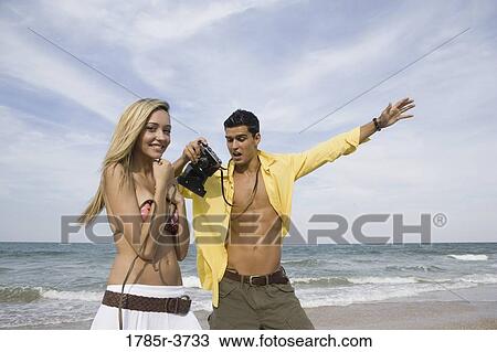 Stock Image - Young man photographing scantily clad woman on the beach. Fotosearch