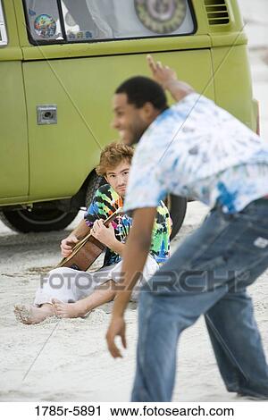 Stock Image - Young man playing guitar by vehicle at beach. Fotosearch