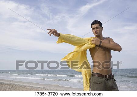Stock Image - Young man putting on a shirt at the beach. Fotosearch