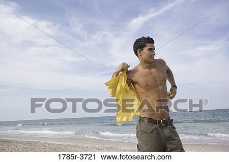 Young man putting on a shirt at the beach View Large Photo Image Stock Image - Young man putting on a shirt at the beach. Fotosearch