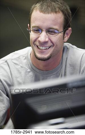 Young man sitting in front of a computer monitor and smiling View Large Photo Image Stock Image - Young man sitting in front of a computer monitor and smiling. Fotosearch
