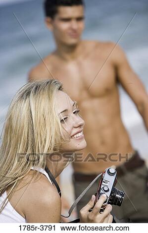 Stock Image - Young woman holding camera with man in background. Fotosearch