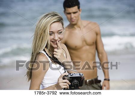 Young woman holding camera with man in background View Large Photo Image Stock Image - Young woman holding camera with man in background. Fotosearch