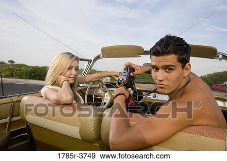 Stock Photo - Young woman sitting in antique convertible car being photographed by a man. Fotosearch