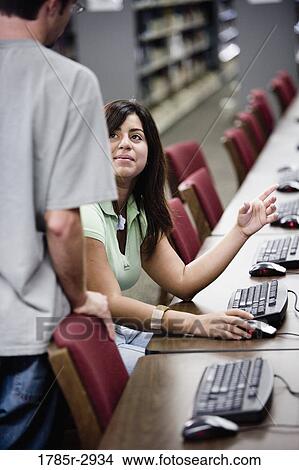 Picture - Young woman using a computer in the library conversing with another student. Fotosearch