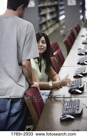 Stock Image - Young woman using a computer in the library conversing with another student. Fotosearch