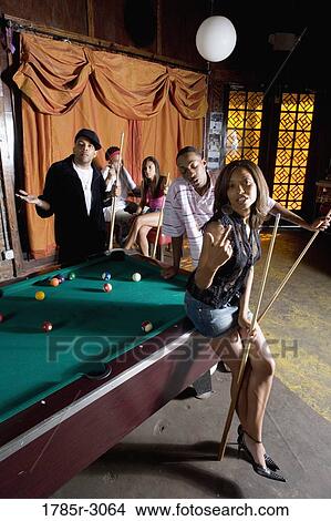 Young woman with pool stick next to pool table with friends watching in background View Large Photo Image Picture - Young woman with pool stick next to pool table with friends watching in background. Fotosearch