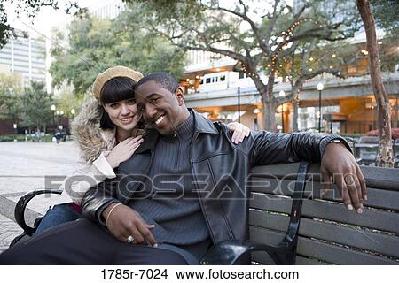Cheerful young inter-racial couple sitting on a park bench View Large Photo Image Picture - Cheerful young inter-racial couple sitting on a park bench. Fotosearch