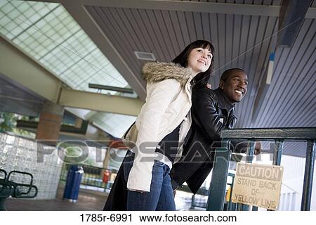 Stock Image - Cheerful young inter-racial couple waiting at the platform of a public train station. Fotosearch
