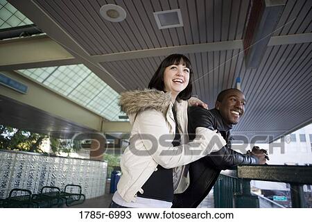 Stock Image - Cheerful young inter-racial couple waiting at the platform of a public train station. Fotosearch