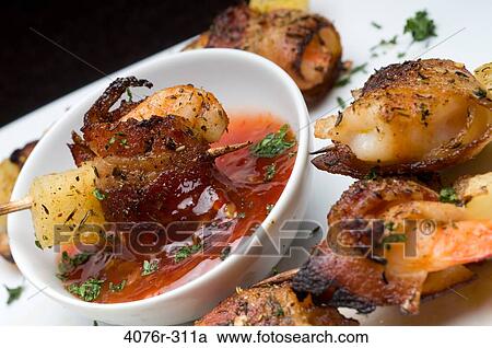 Stock Image - Close-up of bacon wrapped shrimp and pineapple on skewer with a bowl of sauce. Fotosearch