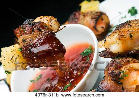 Stock Image - Close-up of bacon wrapped shrimp and pineapple on skewer with a bowl of sauce. Fotosearch