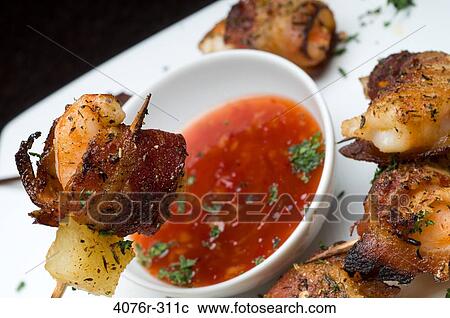 Stock Image - Close-up of bacon wrapped shrimp and pineapple on skewer with a bowl of sauce. Fotosearch