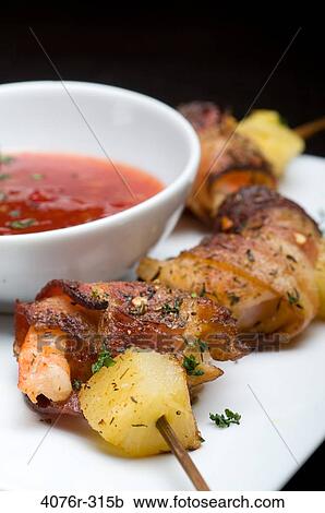 Stock Image - Close-up of bacon wrapped shrimp and pineapple on skewer with a bowl of sauce. Fotosearch