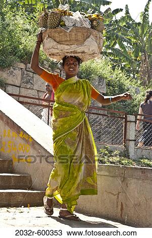 Stock Image - Female fruit seller carrying a basket of pineapples on head, Hampi, Karnataka, India. Fotosearch