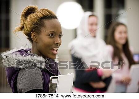 Picture - Head-shot of a young woman holding a book, while other women stand in the background. Fotosearch