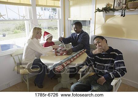 Inter-racial family at kitchen table wrapping gifts while son sits in foreground View Large Photo Image Stock Photograph - Inter-racial family at kitchen table wrapping gifts while son sits in foreground. Fotosearch