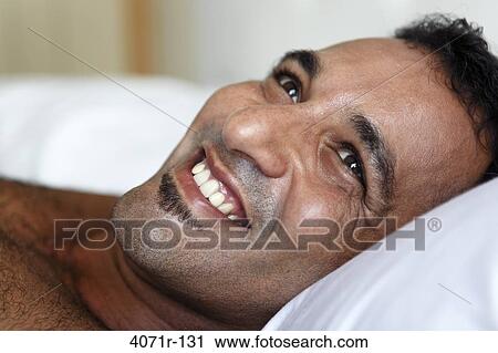 Stock Image - Latin man in bed with head on pillow, looking up, smiling. Fotosearch