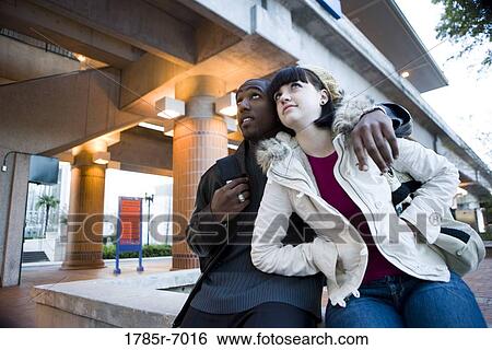Low angle view of a young inter-racial couple sitting on a wall outside a train station View Large Photo Image Stock Photograph - Low angle view of a young inter-racial couple sitting on a wall outside a train station. Fotosearch