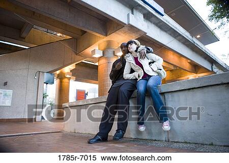 Stock Photography - Low angle view of a young inter-racial couple sitting on a wall outside a train station. Fotosearch