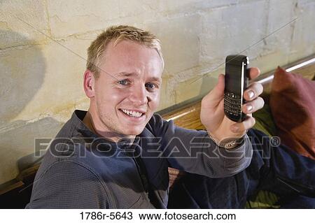Man relaxing on sofa holding up mobile phone View Large Photo Image Stock Image - Man relaxing on sofa holding up mobile phone. Fotosearch