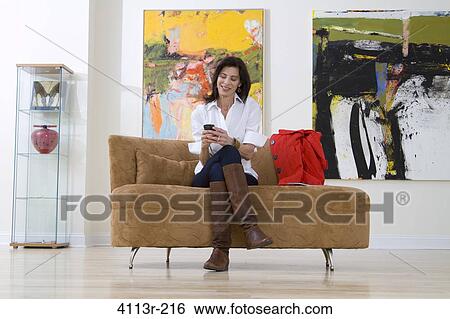 Stock Photograph - Mature woman sitting on sofa and text messaging in art gallery. Fotosearch