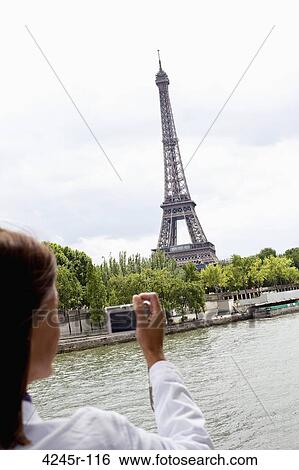 Stock Photograph - Mature woman taking a picture of a tower, Eiffel Tower, Seine River, Paris, Ile-de-France, France. Fotosearch