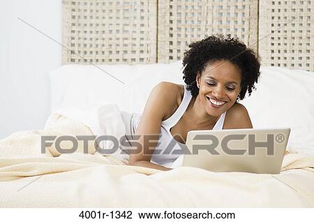 Stock Image - Mid adult woman lying on the bed and using a laptop. Fotosearch