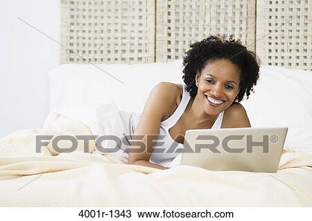 Stock Image - Mid adult woman lying on the bed and using a laptop. Fotosearch