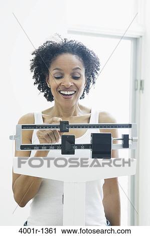 Stock Image - Mid adult woman measuring her weight on a weighing scale. Fotosearch
