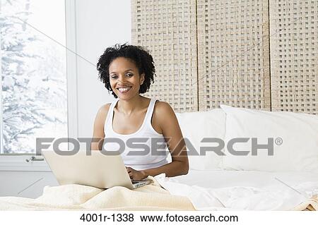 Stock Photo - Mid adult woman sitting on the bed and using a laptop. Fotosearch