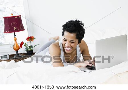 Stock Image - Mid adult woman using a laptop on the bed. Fotosearch