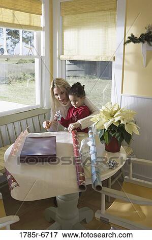 Stock Photo - Mother and young daughter wrapping gifts on kitchen table by large windows. Fotosearch